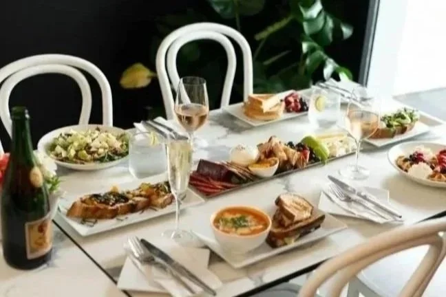 Elegant dining table set with plates of salad, appetizers, bowls of soup, glasses of wine and champagne, and a bottle of champagne, in a well-lit room with white chairs and a window.