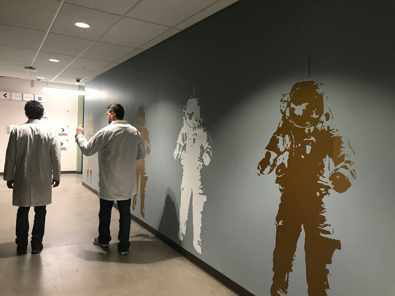 Two people in lab coats walk down a hallway with astronaut silhouettes on the wall.