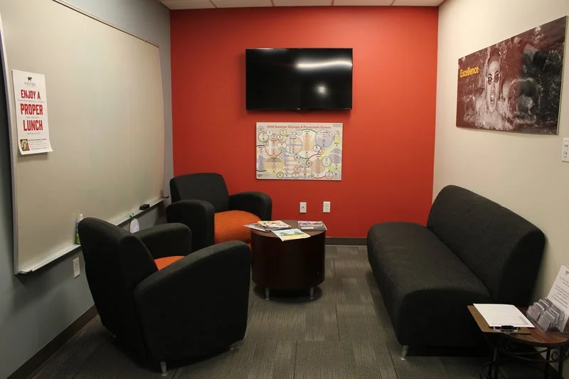 Office lounge with two armchairs, a couch, and a small table, featuring a wall-mounted TV and a whiteboard. The walls display motivational posters.