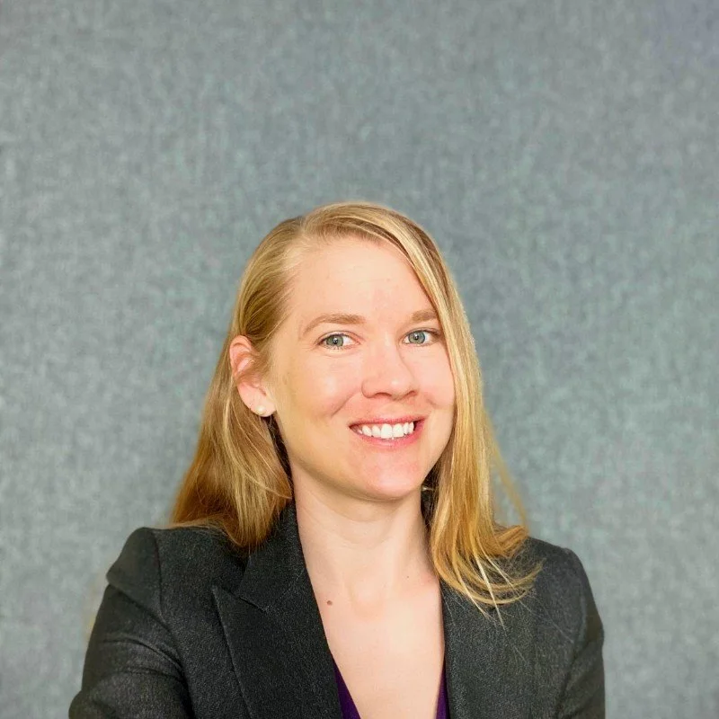 A woman with blonde hair and a black blazer smiling at the camera, standing against a plain, textured gray background.