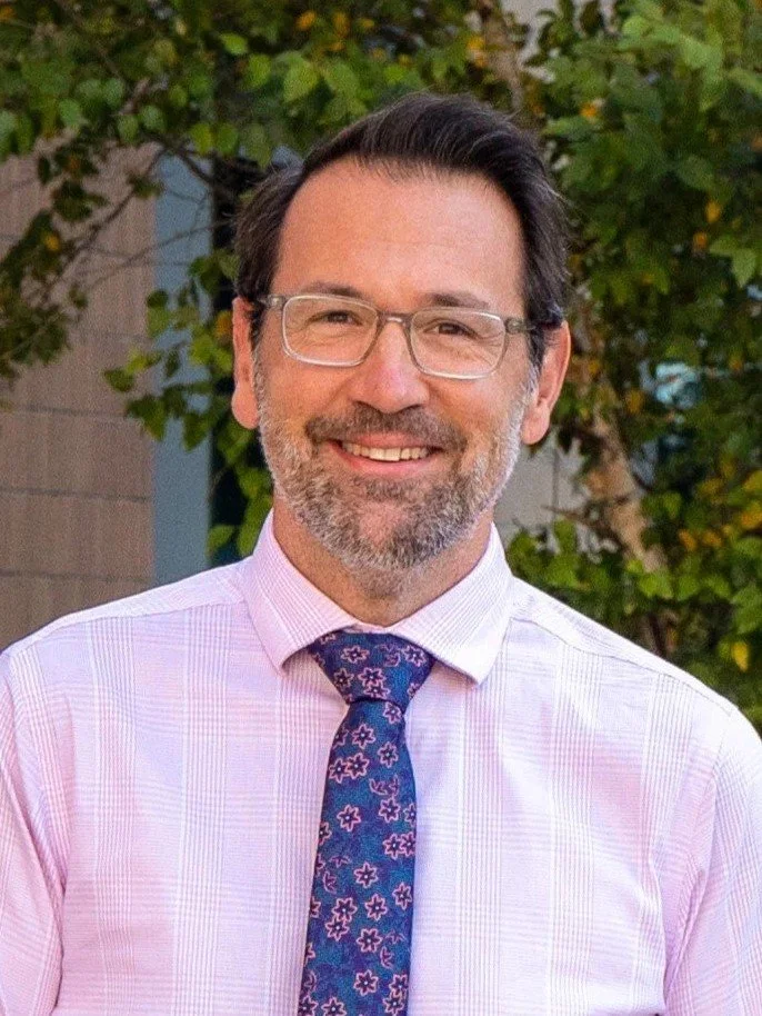 A man with dark hair, glasses, and a beard, wearing a pink checkered dress shirt and a navy blue tie with pink floral patterns, smiling outdoors with trees and a building in the background.