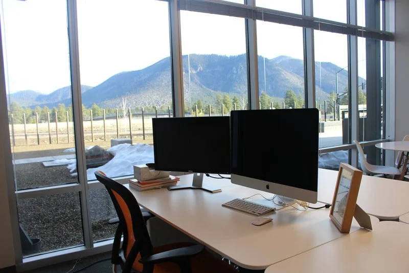Office desk with two computer monitors and a scenic mountain view through large windows.
