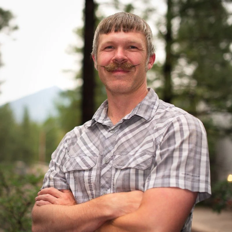 Man with a mustache and short hair smiling outdoors, wearing a plaid shirt, with trees and mountains in the background.