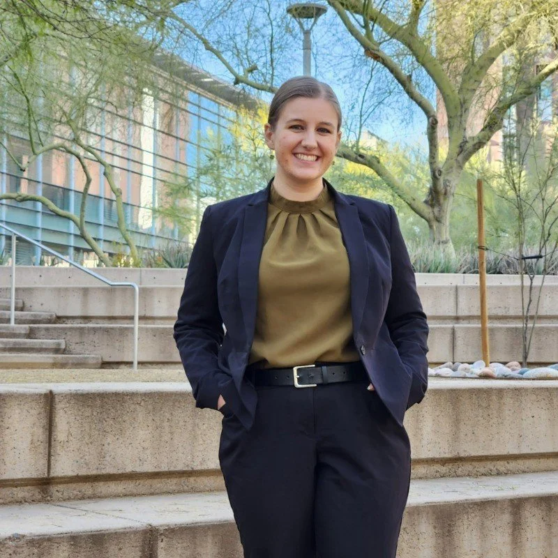 A smiling woman stands outdoors on concrete steps, wearing a navy blazer, khaki blouse, black pants, and a belt, with trees and a building in the background.