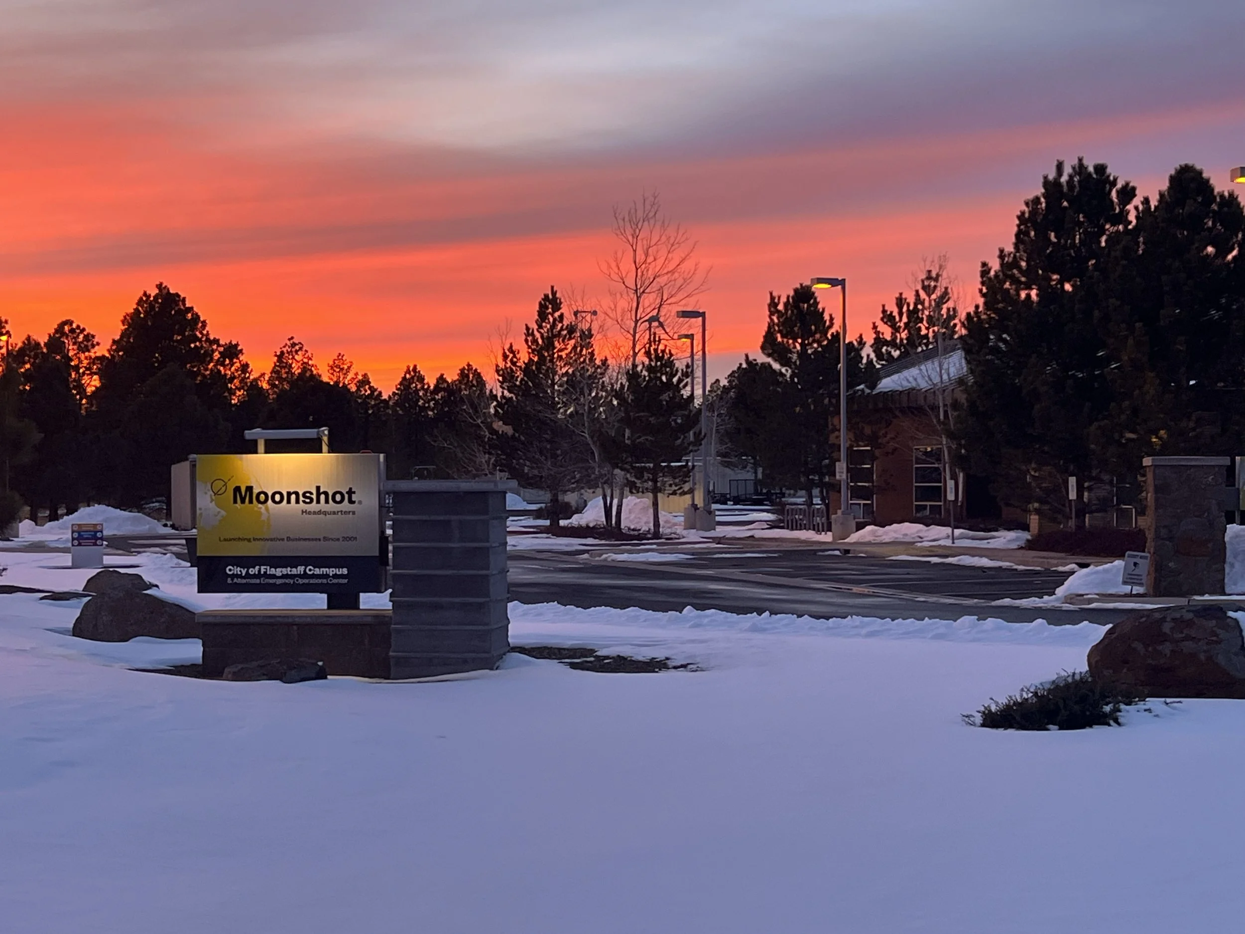 Snow-covered landscape with a sign for Moonshot Headquarters against a vibrant sunset sky, surrounded by trees.
