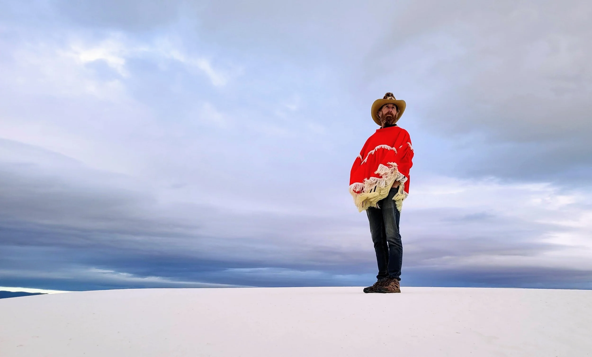 A man wearing a cowboy hat and a red poncho with tassels standing on a white snowy landscape under a cloudy sky.