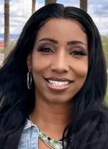 Close-up of a smiling woman with long dark hair, wearing hoop earrings, a denim jacket, and a necklace, outdoors with trees and a cloudy sky in the background.