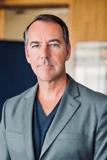 Portrait of a middle-aged man with short dark hair, wearing a gray blazer and navy shirt, standing indoors with a blurred office background.