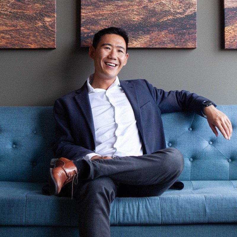 A man in business attire sitting on a blue sofa, smiling, with artwork hanging on a gray wall behind him.