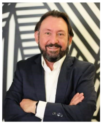 A middle-aged man with dark hair and a beard, wearing a dark suit and white shirt, smiling with his arms crossed in front of a black and white geometric patterned background.