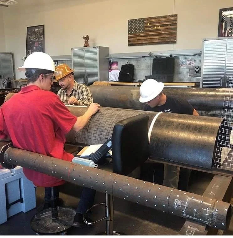 Three construction workers wearing hard hats and casual clothing appear to be attending a meeting or working on a project. They are sitting at a table designed to look like large metal pipes. The room has industrial decor with American-themed elements visible on the wall.