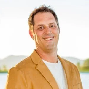 A man smiling outdoors with a blurred nature background, wearing a tan blazer over a white shirt.