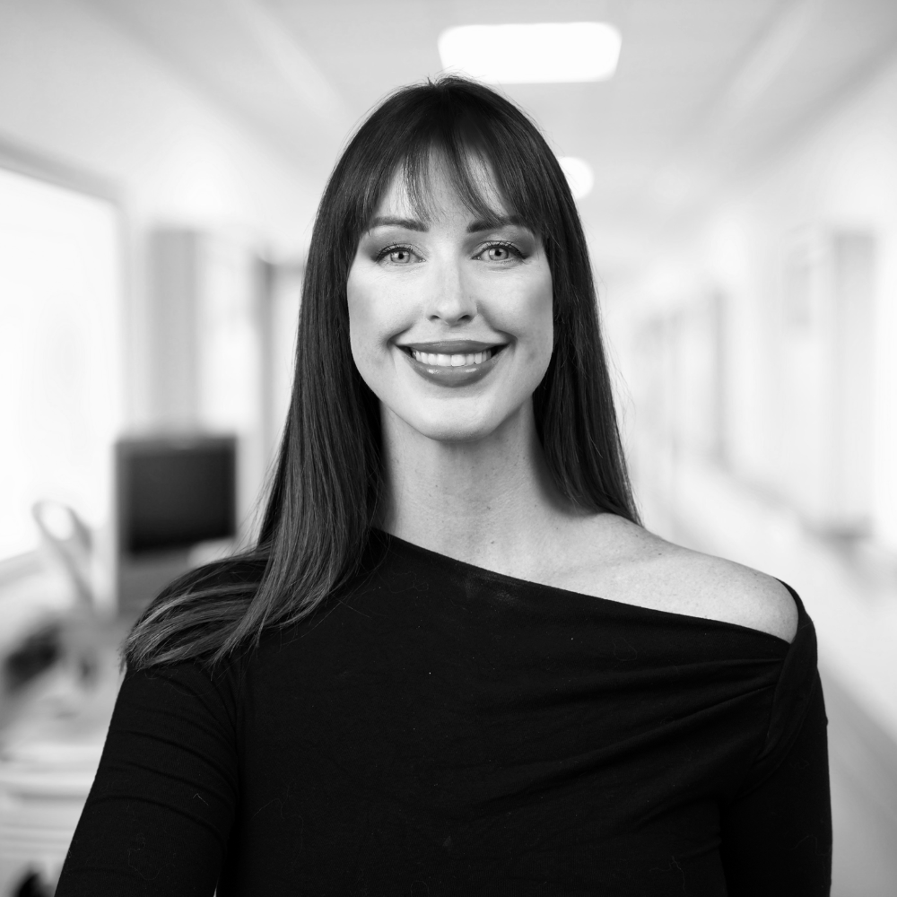A woman with long dark hair and a black off-the-shoulder top, smiling at the camera, in a blurred indoor setting.