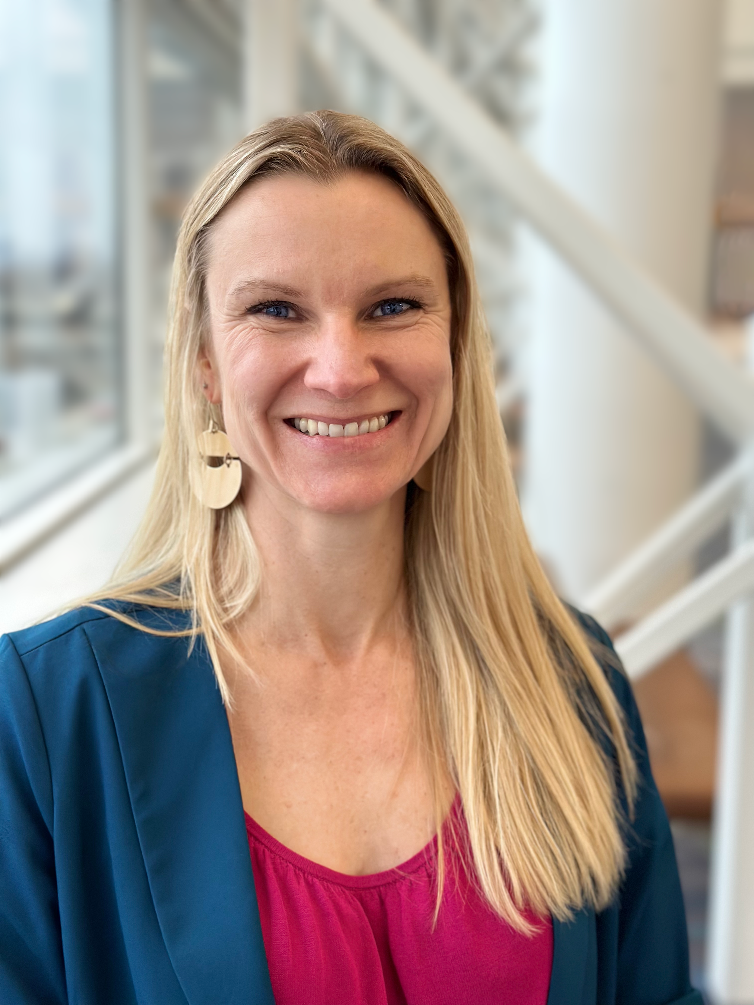 A woman with long blonde hair smiling, wearing a dark blue jacket, pink top, and dangling earrings, standing in front of a staircase near a window.
