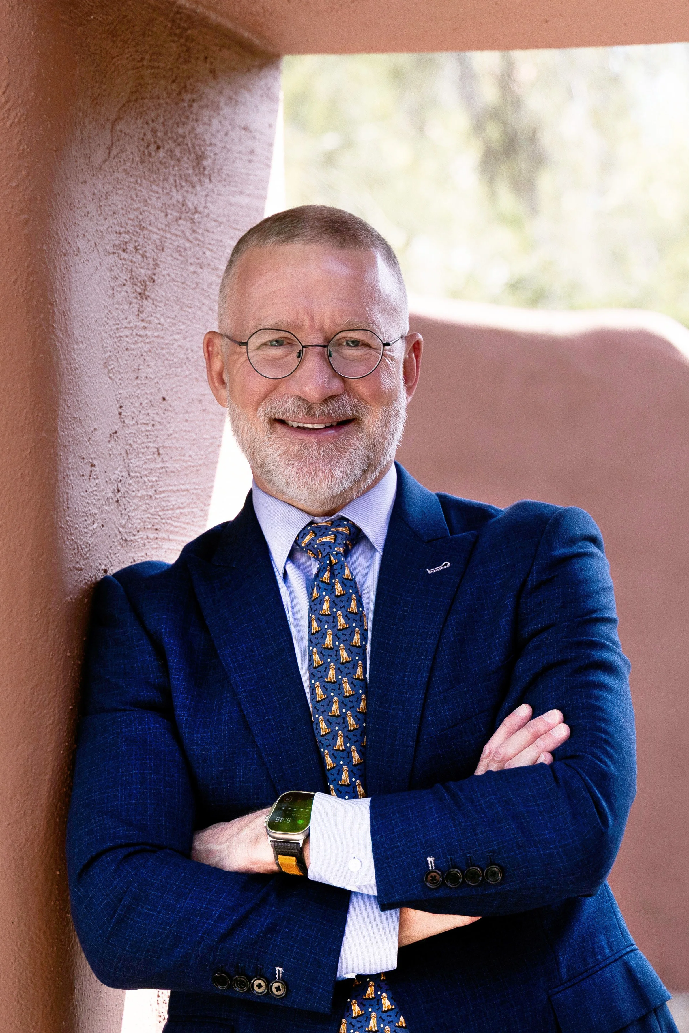 A middle-aged man in a navy suit with a patterned tie, glasses, and a smartwatch, smiling and crossing his arms, standing outdoors against a textured pink wall.