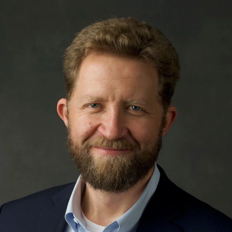 Portrait of a middle-aged man with light brown hair and a beard, wearing a dark suit and white shirt, smiling against a dark background.