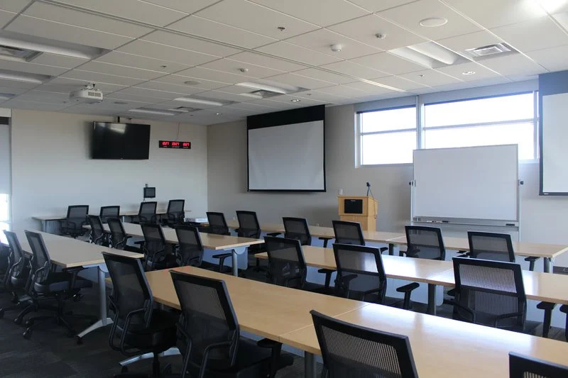 Empty classroom with rows of desks and chairs, a projector screen, whiteboard, wall-mounted clock, and windows.