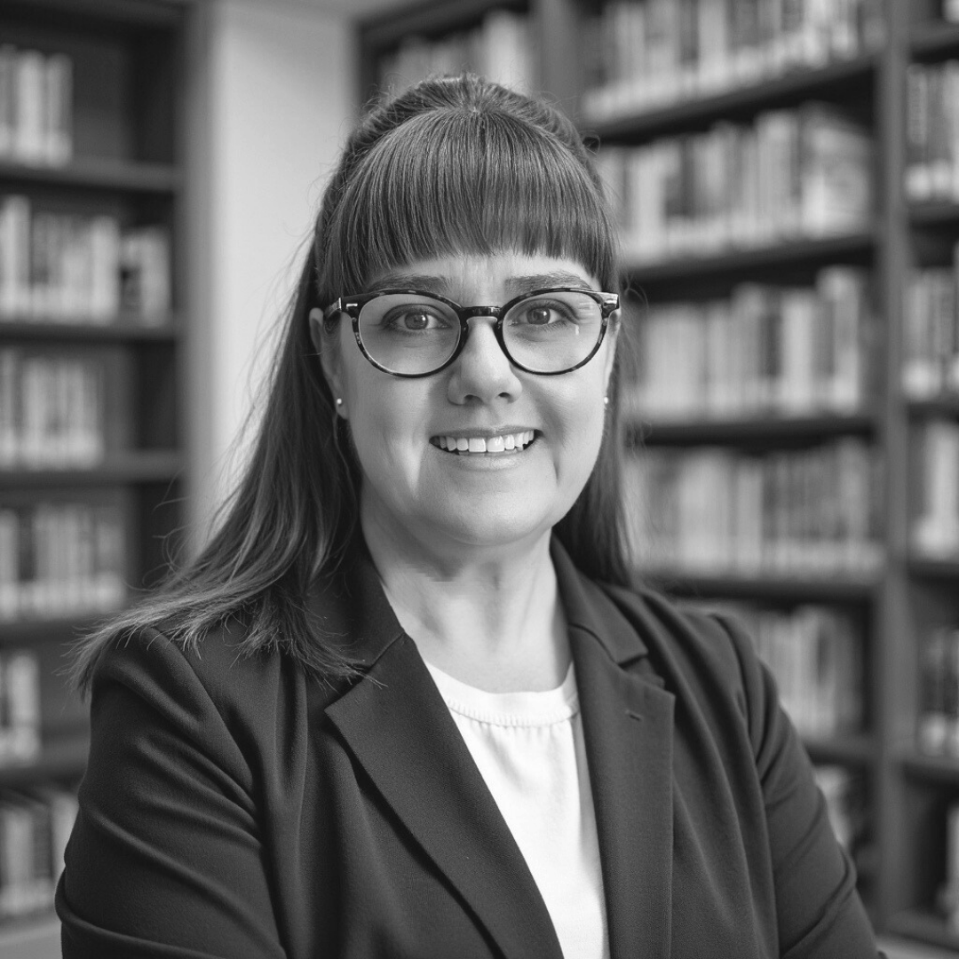 Black and white photo of a person wearing glasses and a blazer, smiling in a library setting.