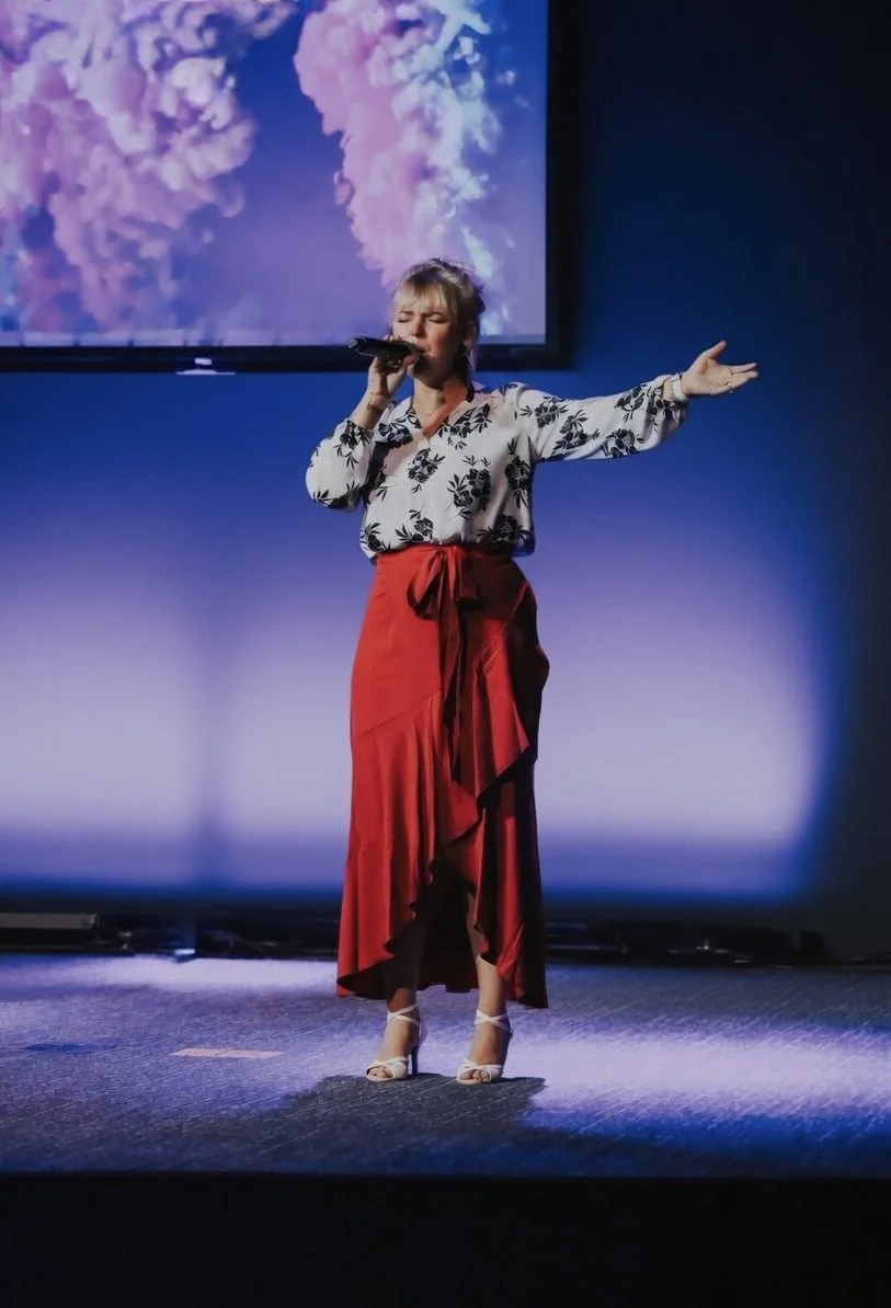A woman singing into a microphone with her eyes closed, standing on a stage with purple lighting and a large screen displaying clouds behind her.