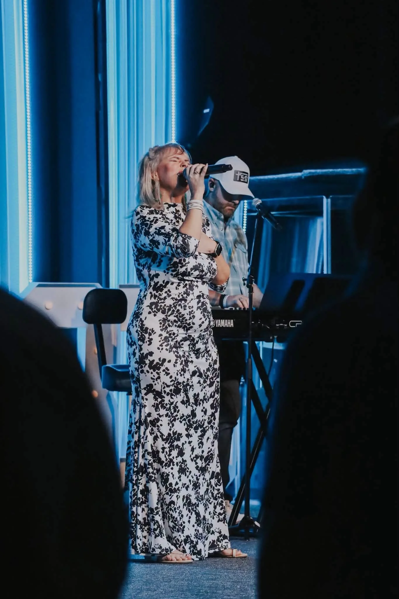 A woman singing into a microphone on stage, dressed in a white and black floral dress, with a man playing keyboard in the background.
