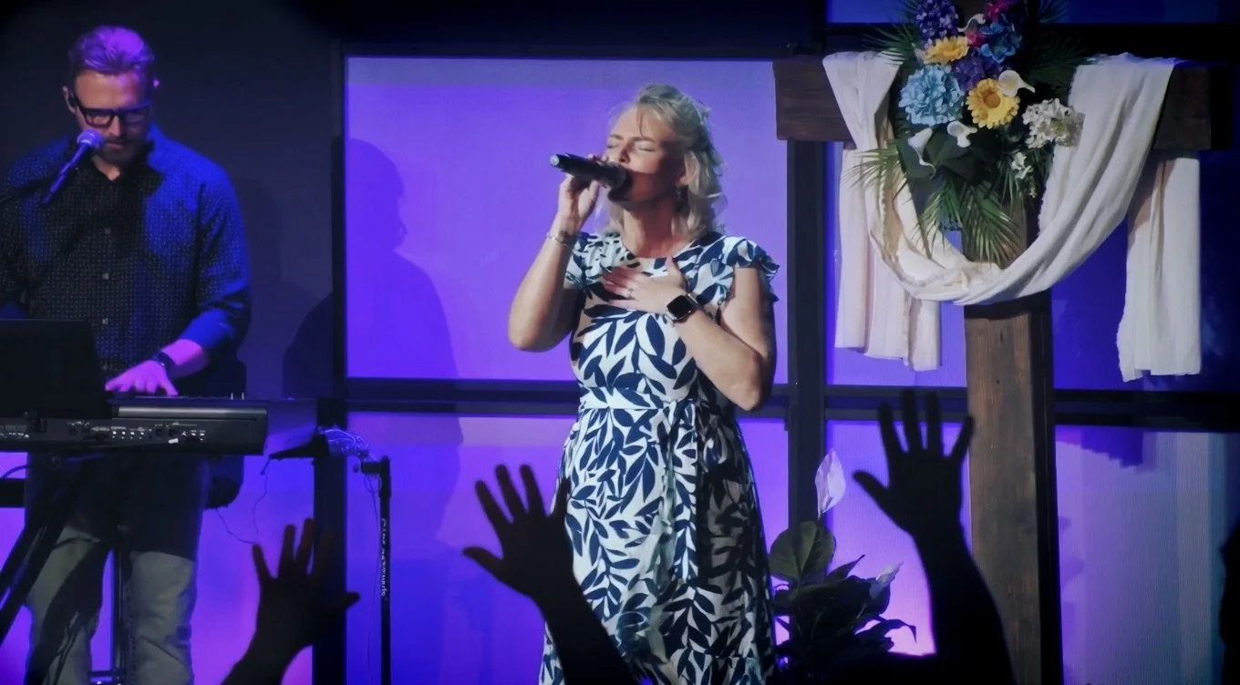 A woman singing into a microphone with her eyes closed, standing in front of a cross decorated with flowers and cloth, with hands raised in the audience. A man is playing a keyboard next to her. The setting appears to be a worship service or concert 
