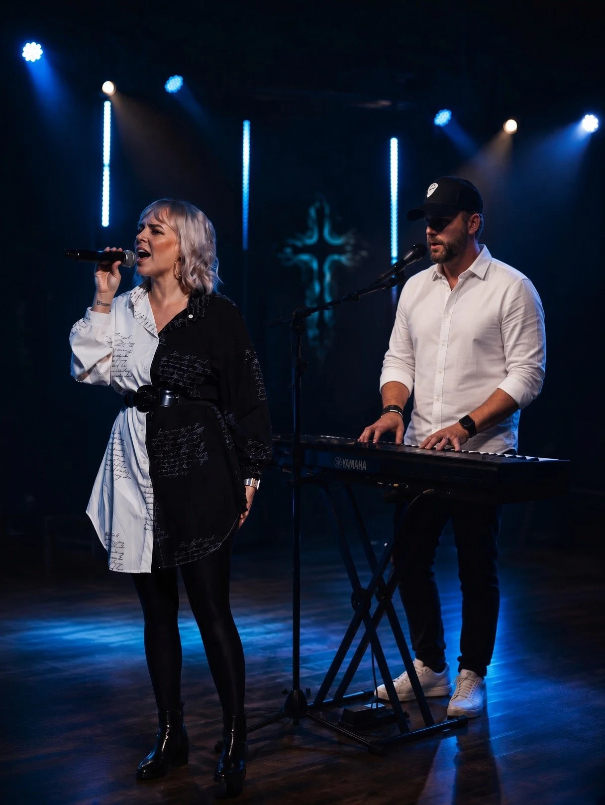 A woman singing into a microphone and a man playing a keyboard on stage with blue lighting and a cross symbol in the background.