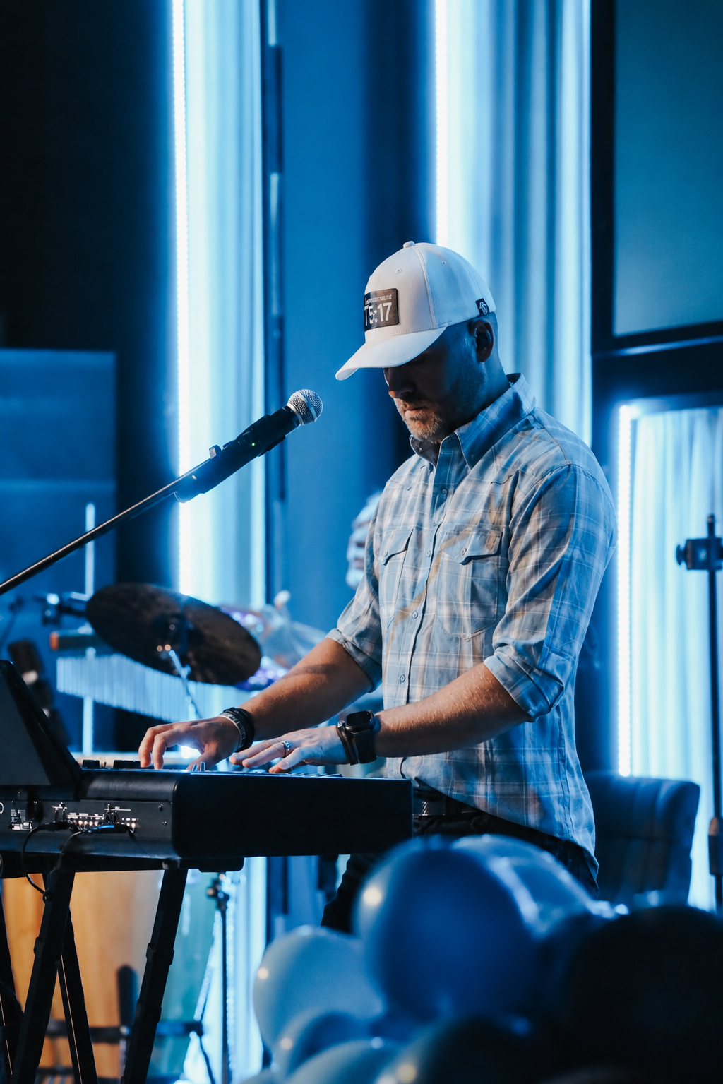 A man wearing a white baseball cap and plaid shirt playing keyboard on stage with blue lighting and balloons in the foreground.