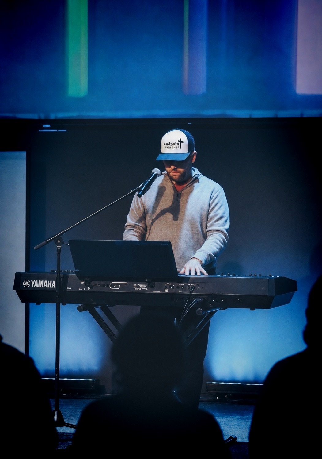 Man playing a Yamaha keyboard on stage, wearing a gray hoodie and a baseball cap with 'endpoints worship' written on it, in front of a microphone.