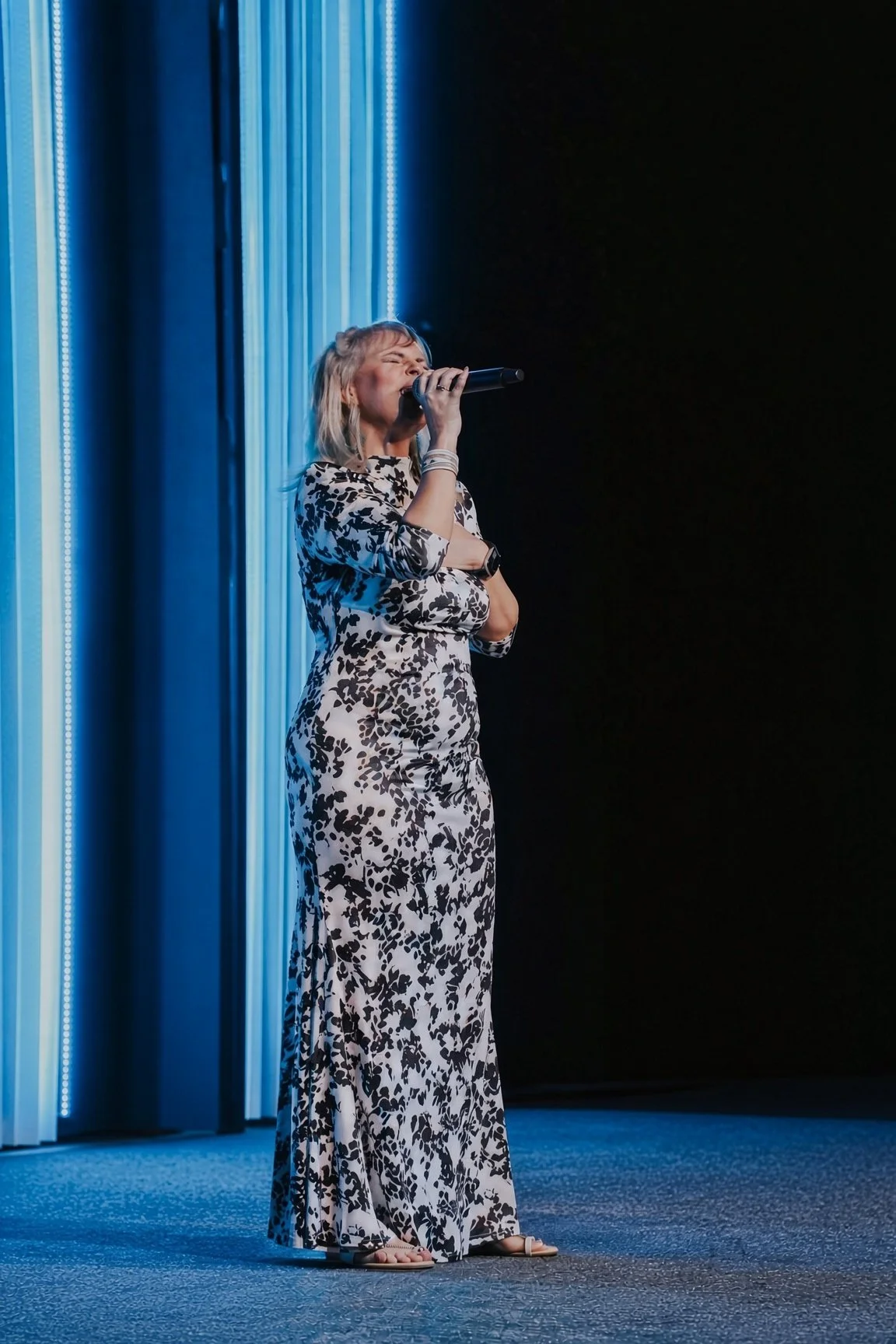 A woman with blonde hair singing into a microphone on stage, wearing a long, black and white floral dress, with blue curtains and stage lighting in the background.