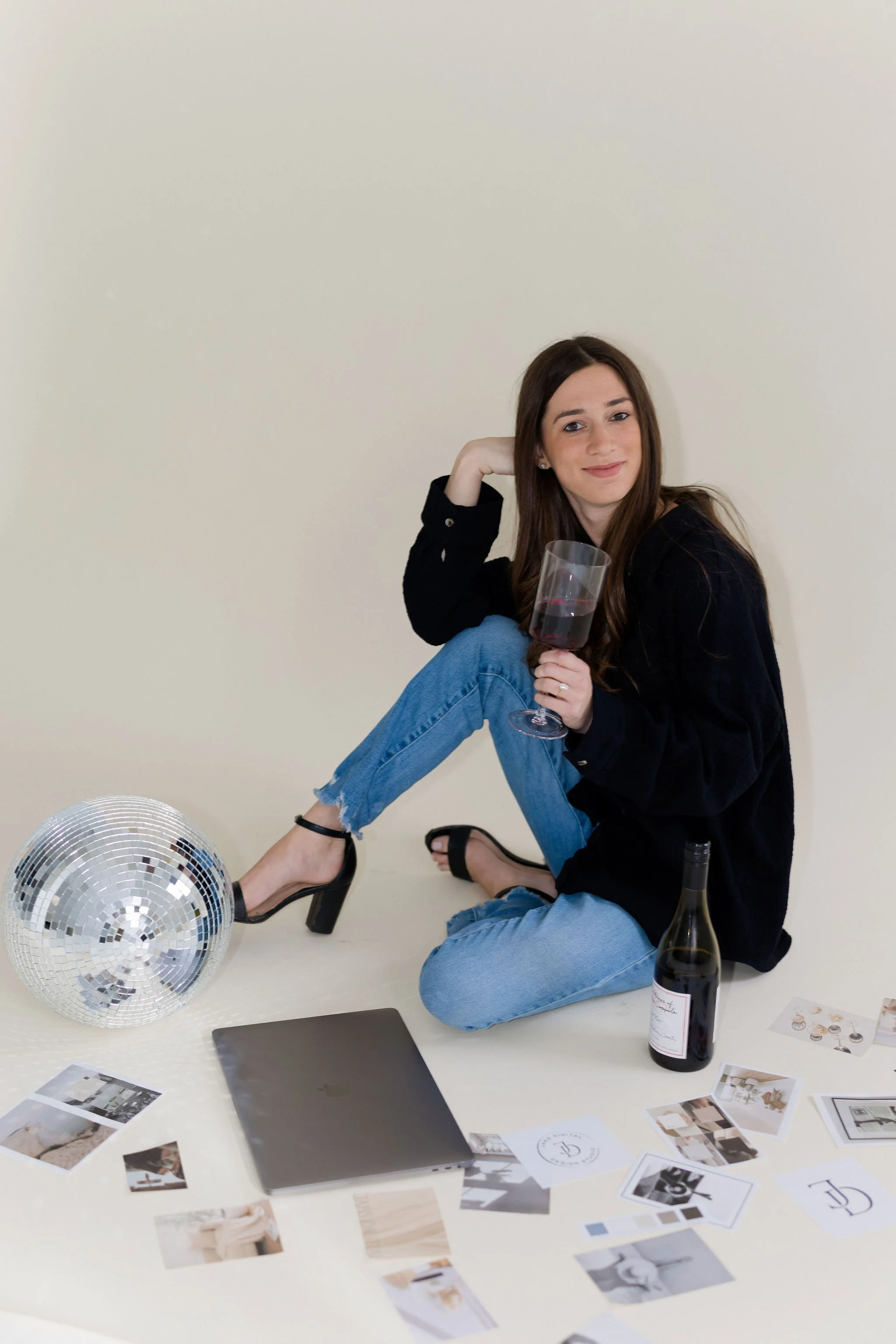 A young woman with long brown hair sitting on the floor, holding a glass of red wine, surrounded by a disco ball, a bottle of wine, a closed laptop, and scattered photographs and papers, against a plain light-colored wall.