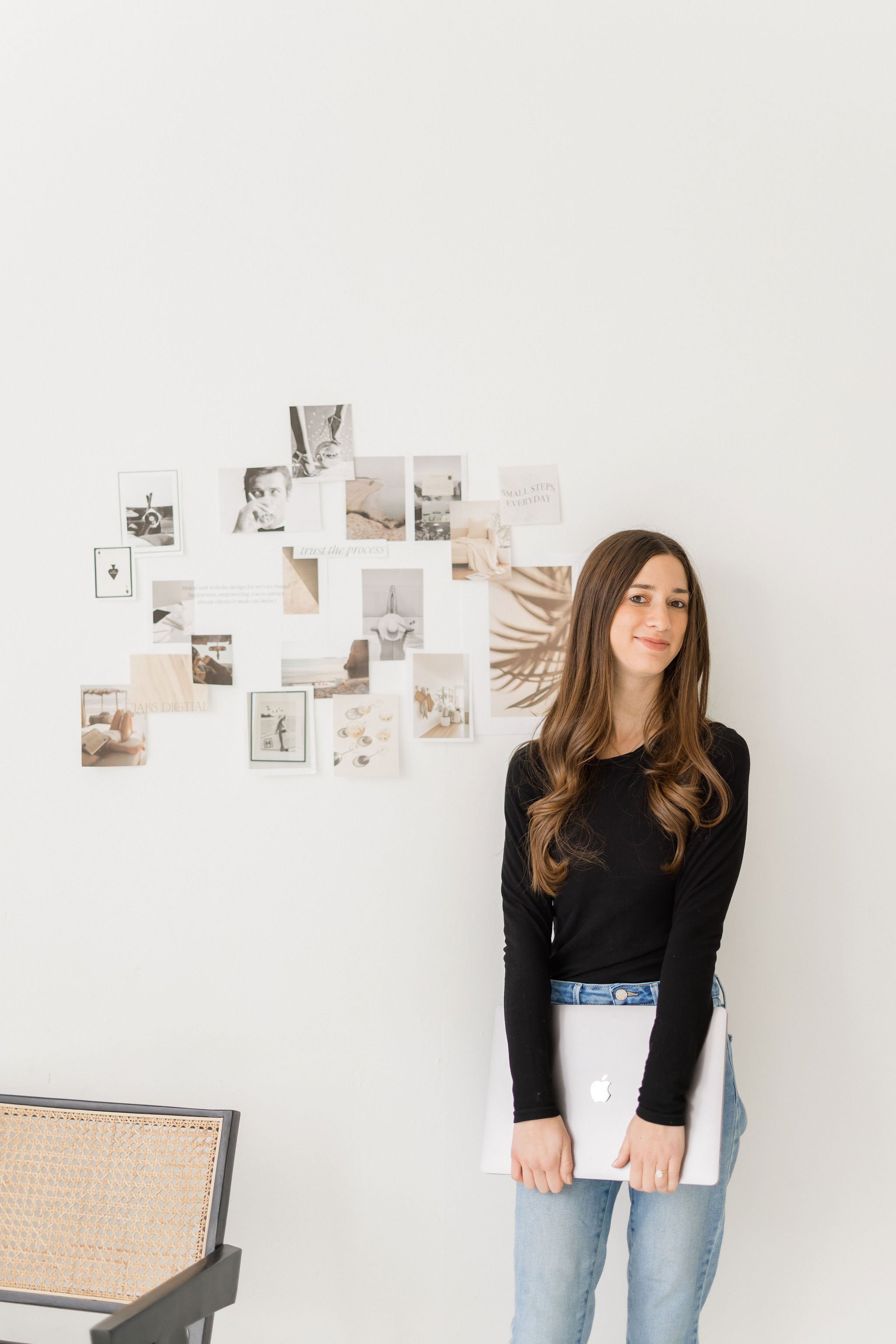 A young woman with long brown hair, wearing a black long-sleeve shirt and light blue jeans, holding a closed MacBook laptop in front of her, standing in a room with white walls and a collage of photographs and images on the wall behind her.