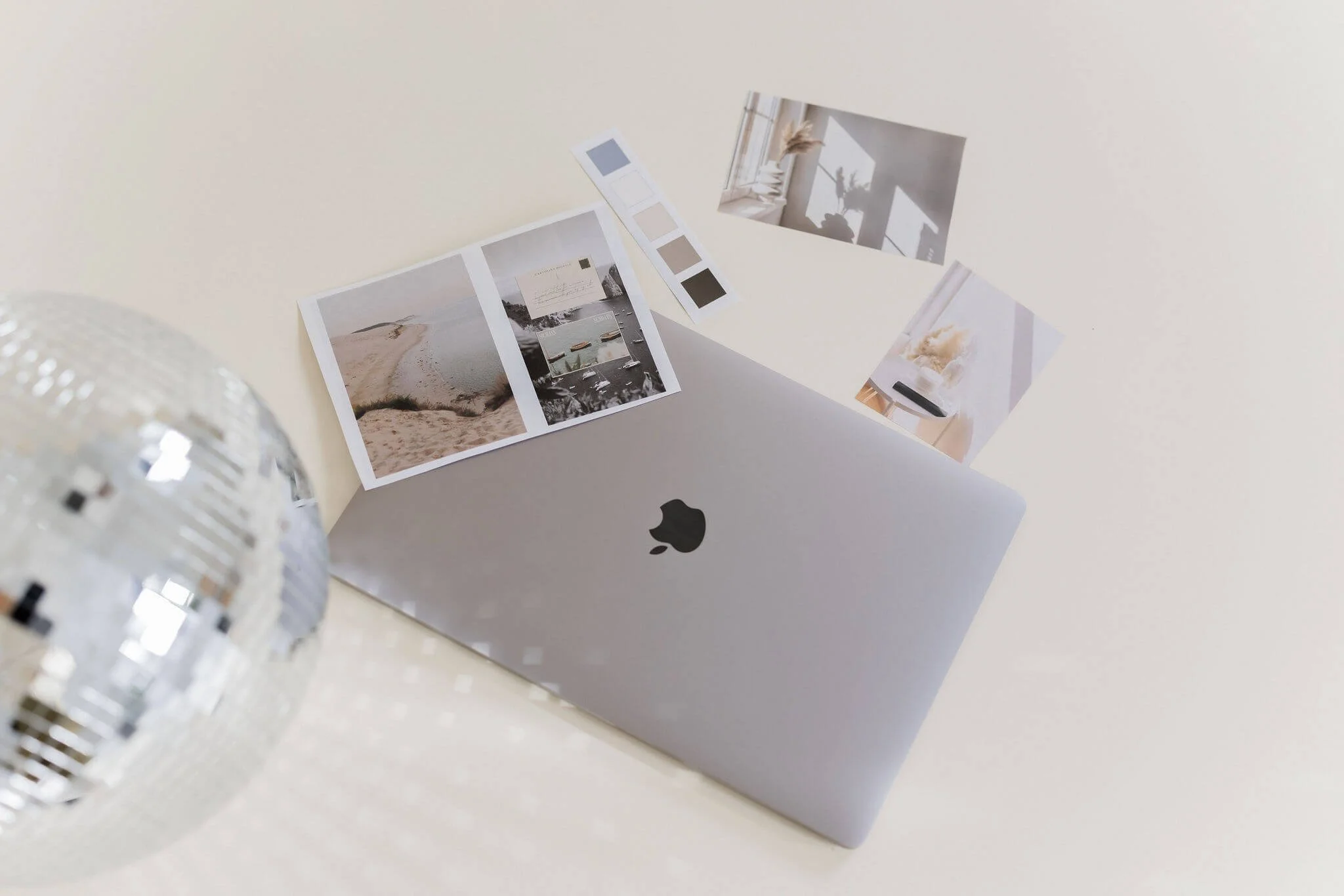Open silver MacBook laptop with photos and color palette papers around it on a white surface, with a mirrored disco ball on the left.