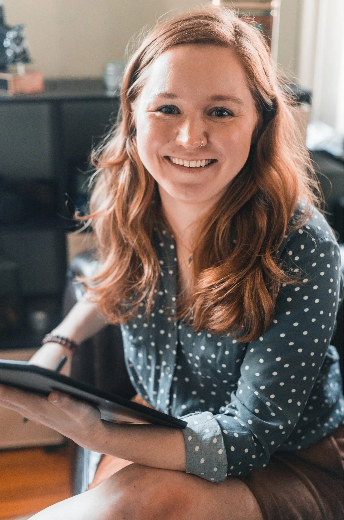 A woman with long wavy red hair smiling and holding a tablet in a cozy indoor setting.