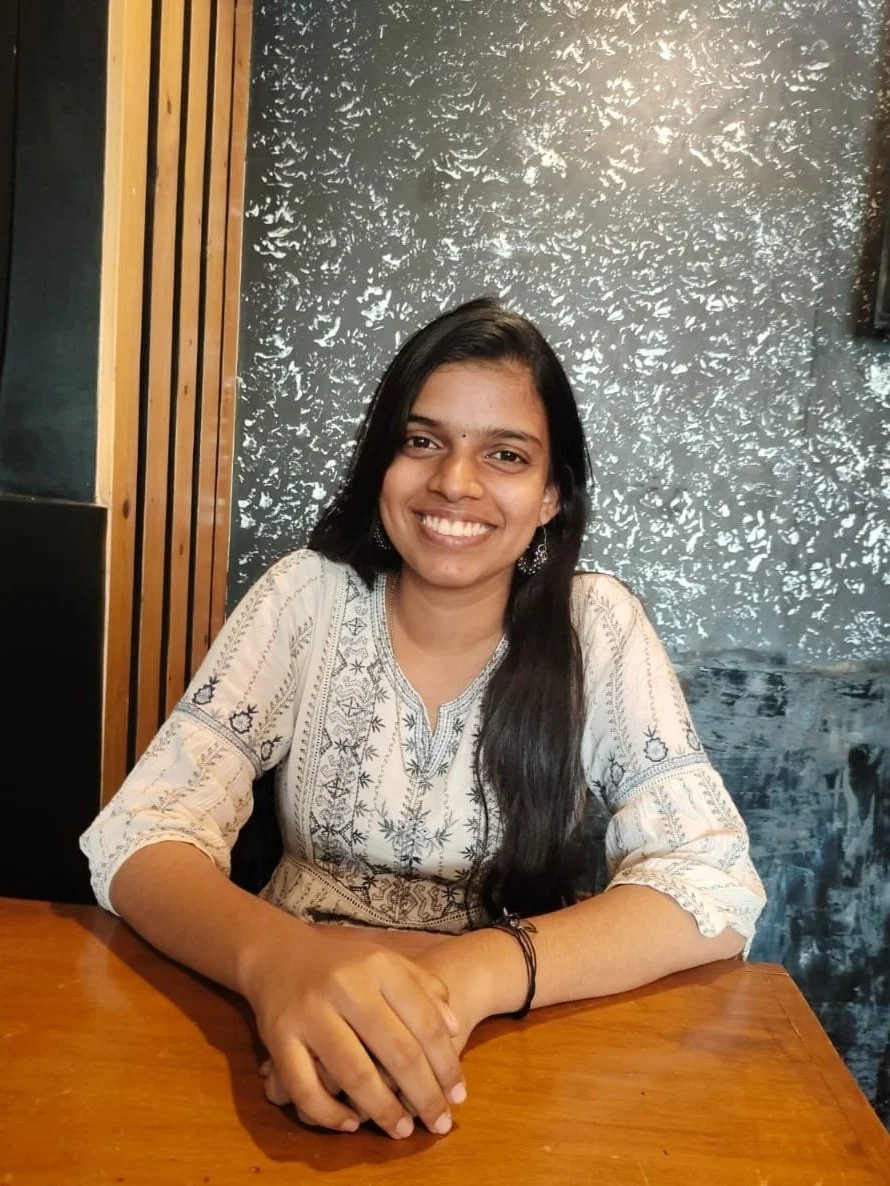 A smiling woman with long black hair sitting at a wooden table in a restaurant or cafe.