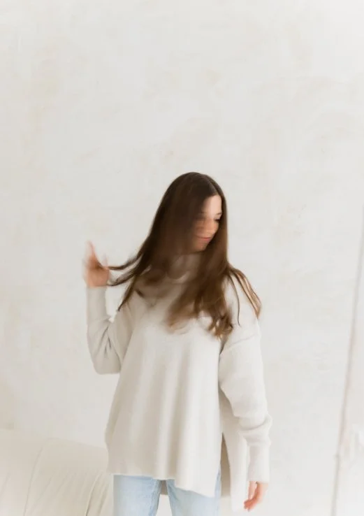A woman with long brown hair wearing a beige sweater and light blue jeans, standing against a plain white wall.