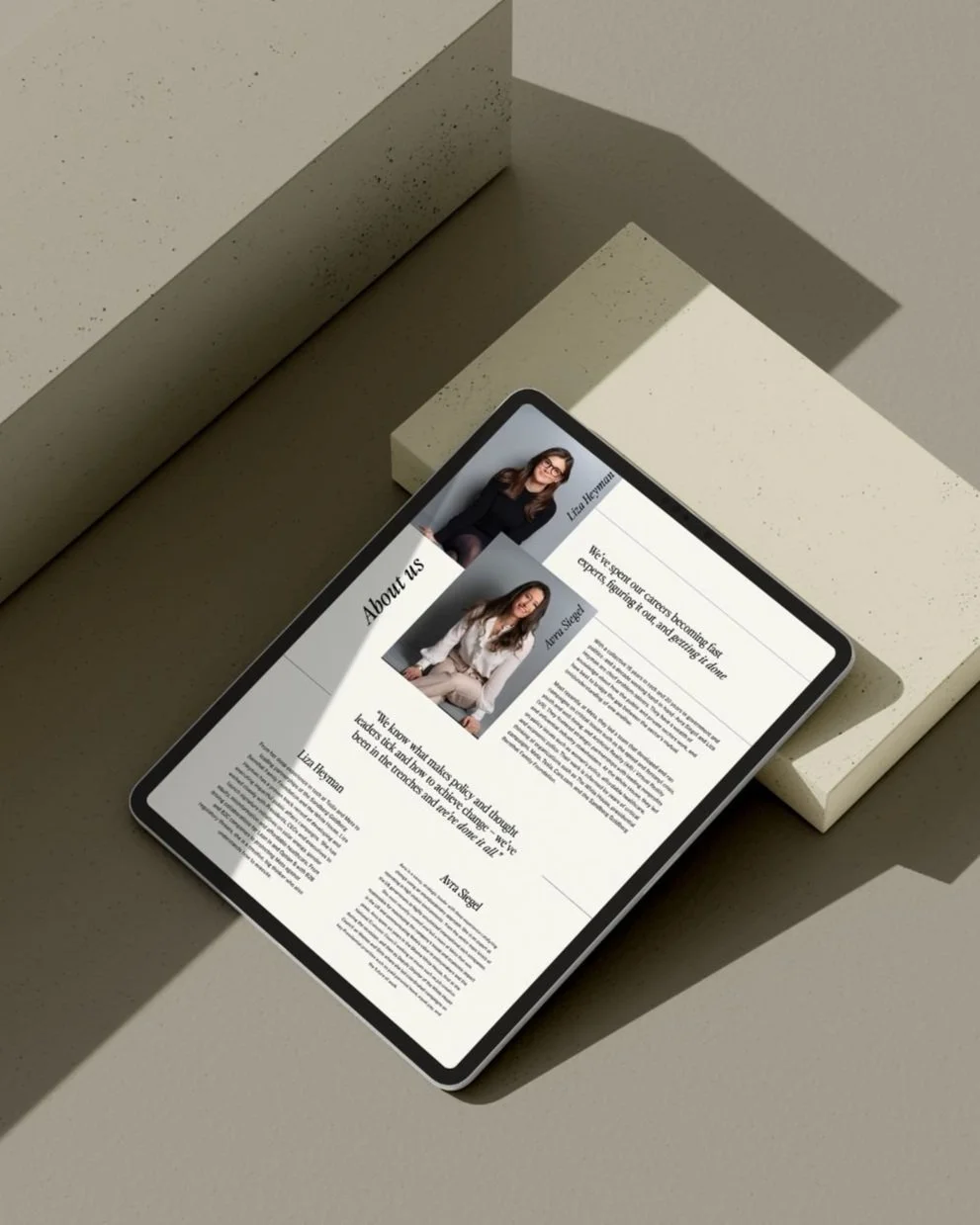 A tablet displaying an 'About Us' webpage with photos and bios of three women, placed on a neutral-colored surface with geometric blocks around it.