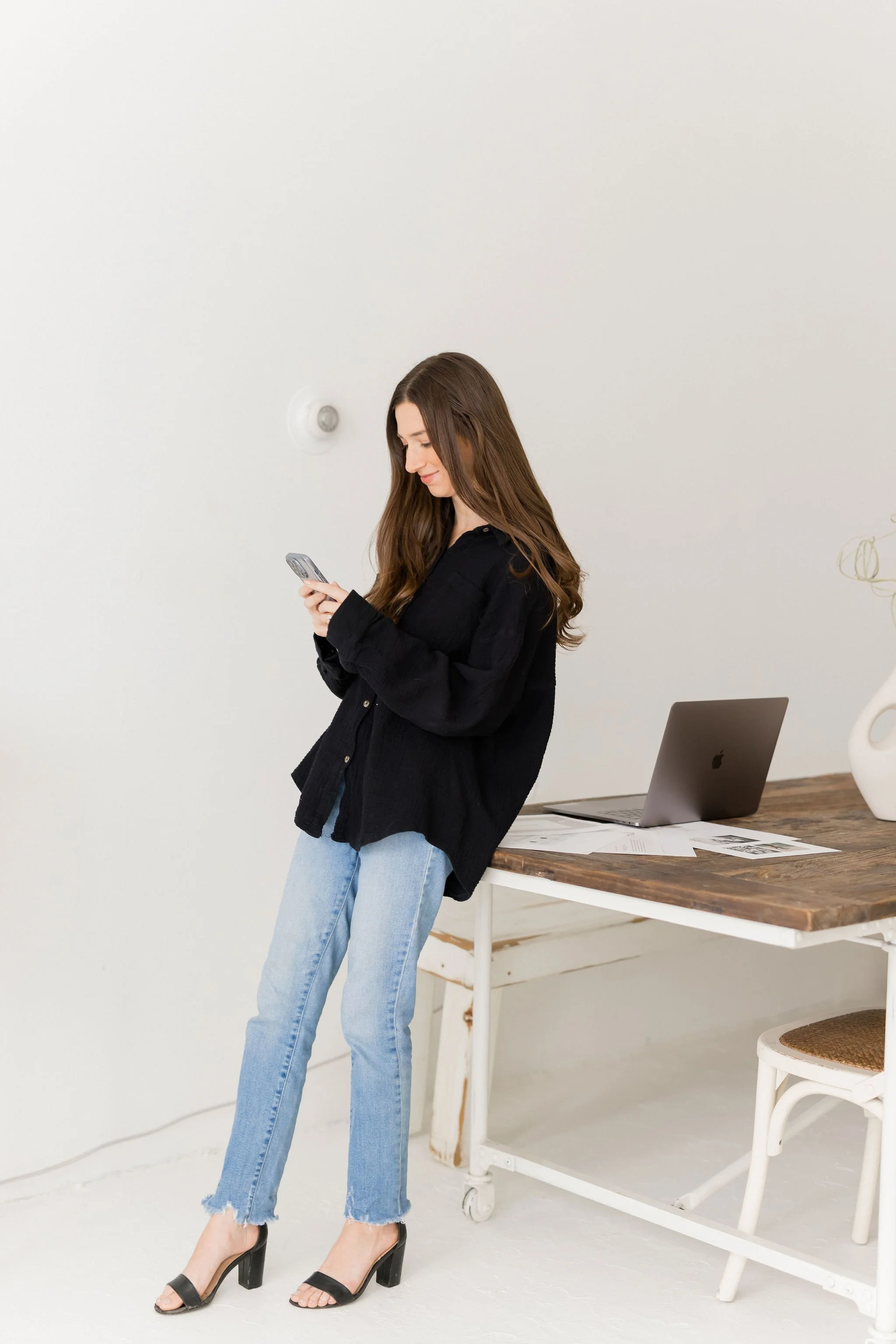 Young woman in black shirt and jeans looking at her phone in a bright, minimalist office with a wooden table, a laptop, papers, and a white vase.