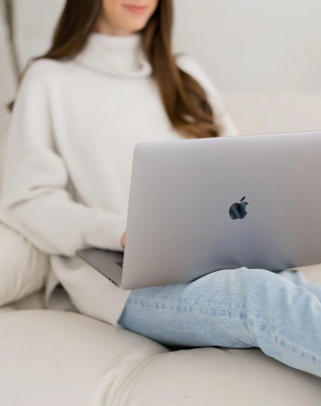 Woman in white sweater and light blue jeans sitting on a beige sofa using a silver MacBook laptop.