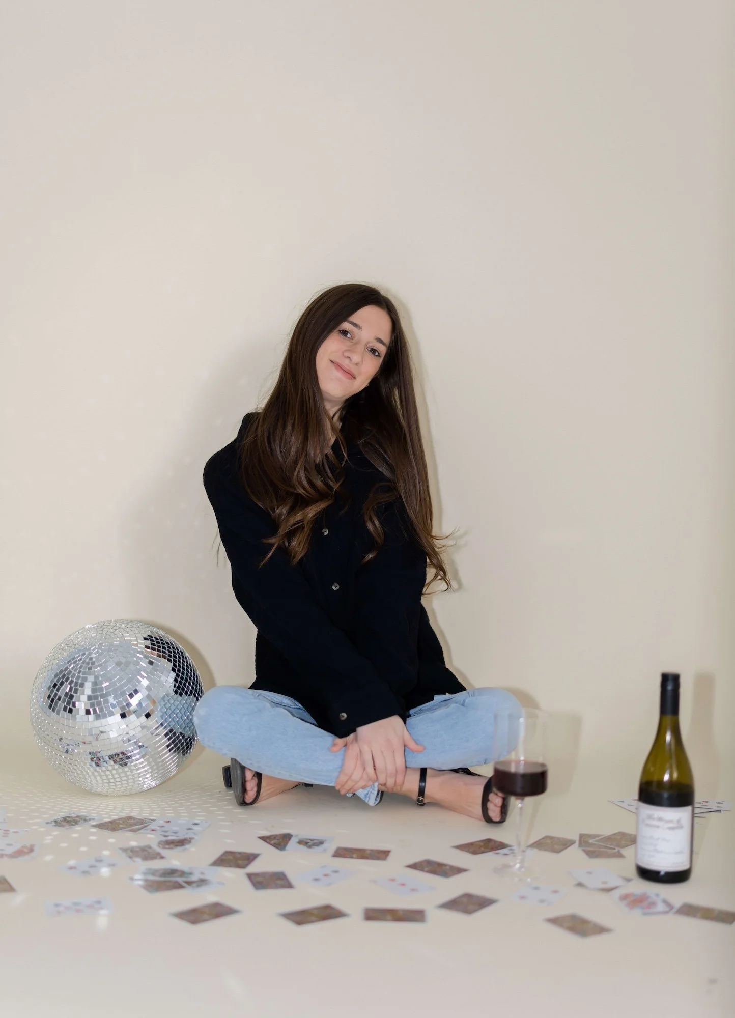 A young woman sitting on the floor with crossed legs, wearing a black jacket, light blue jeans, and sandals, surrounded by a disco ball, scattered playing cards, a glass of red wine, and a bottle of wine against a plain beige wall.