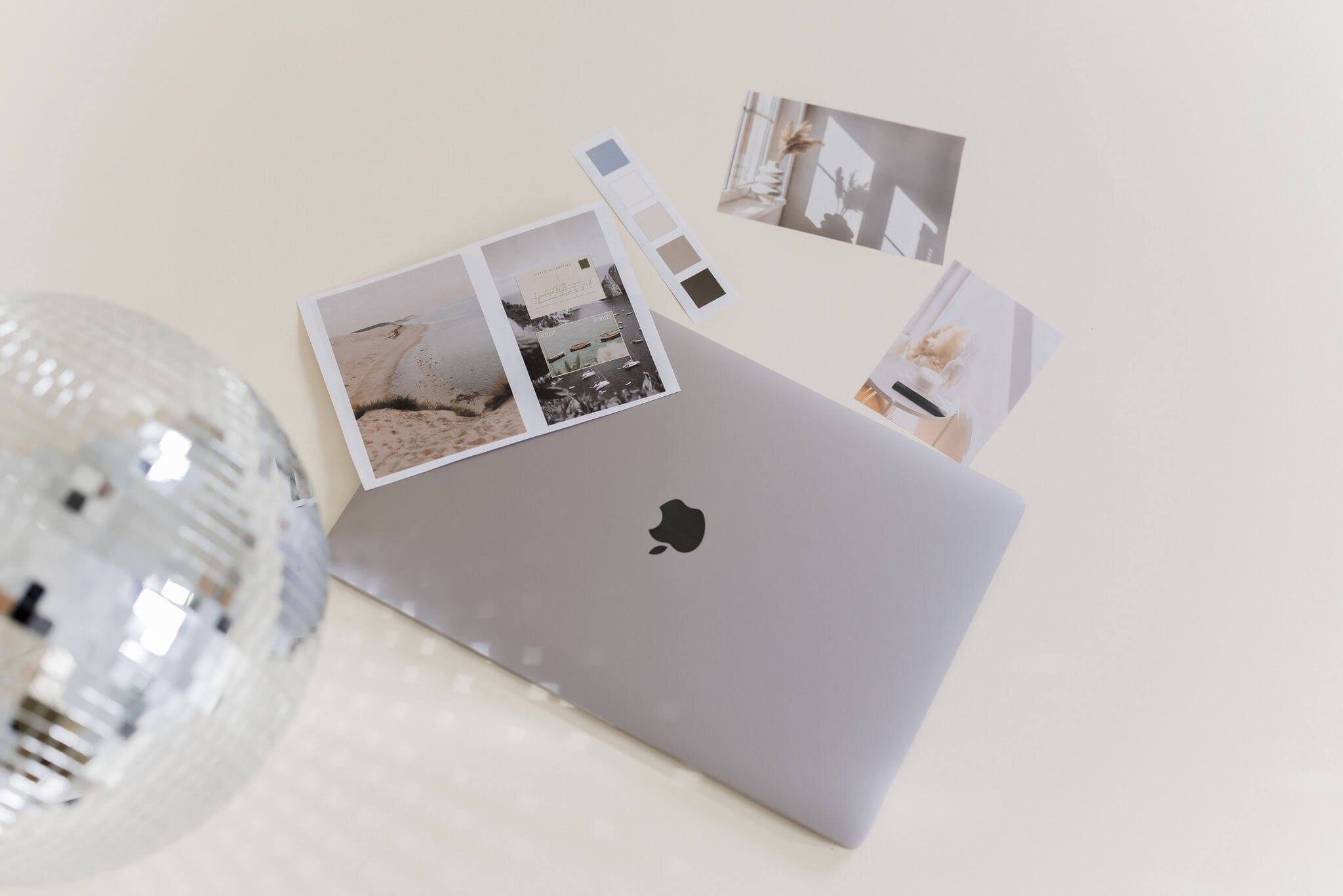 A closed MacBook with photographs and color swatches spread around it on a white surface, a blurred disco ball on the left side.