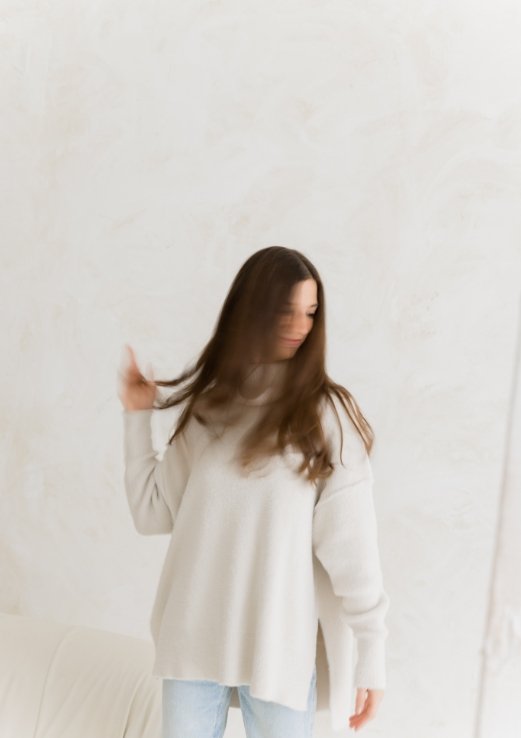 A woman with long brown hair wearing a white sweater and light blue jeans standing against a plain white wall.