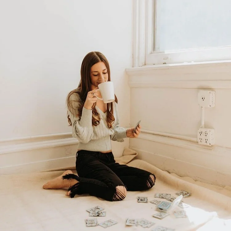 A young woman sitting on the floor by a window, drinking from a large mug and holding a phone, surrounded by scattered playing cards.