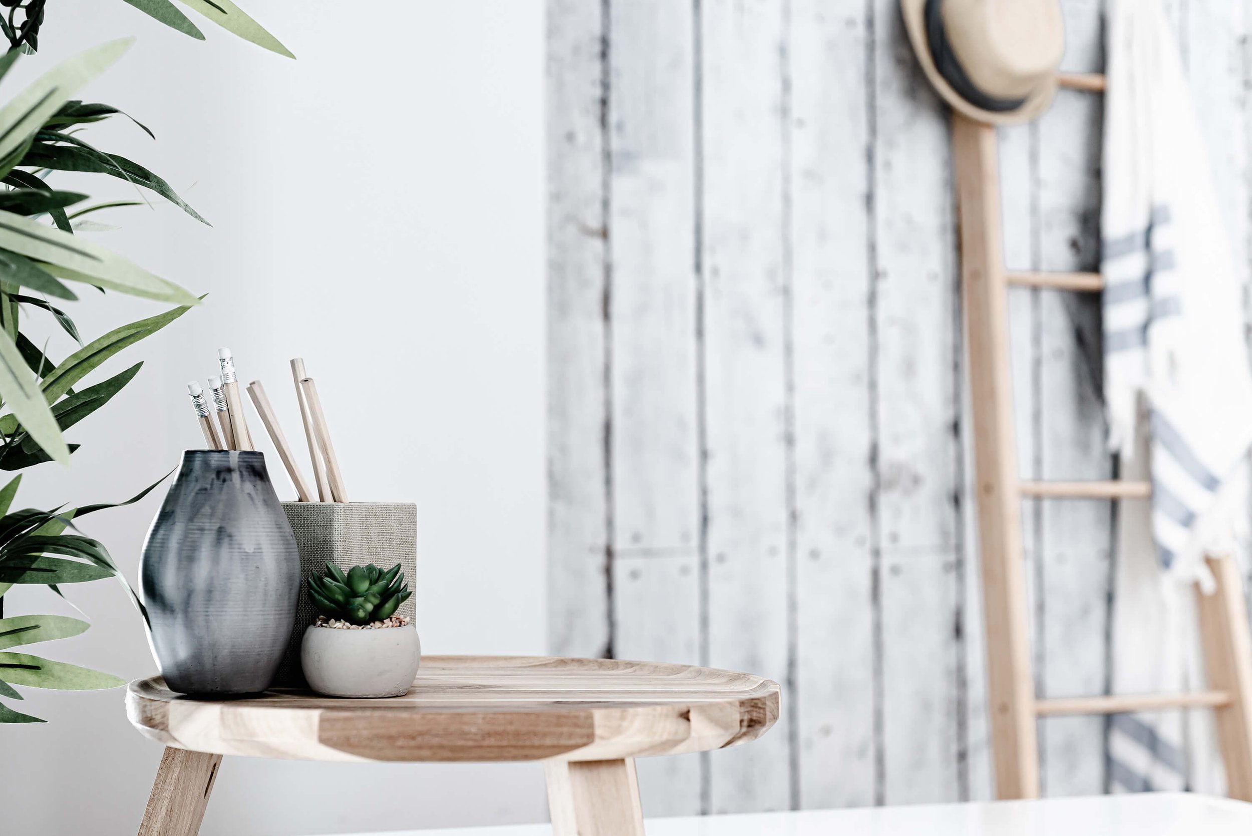 Decorative table with a vase, a container with pencils, and a small succulent plant, against a rustic wooden wall and ladder with hats.