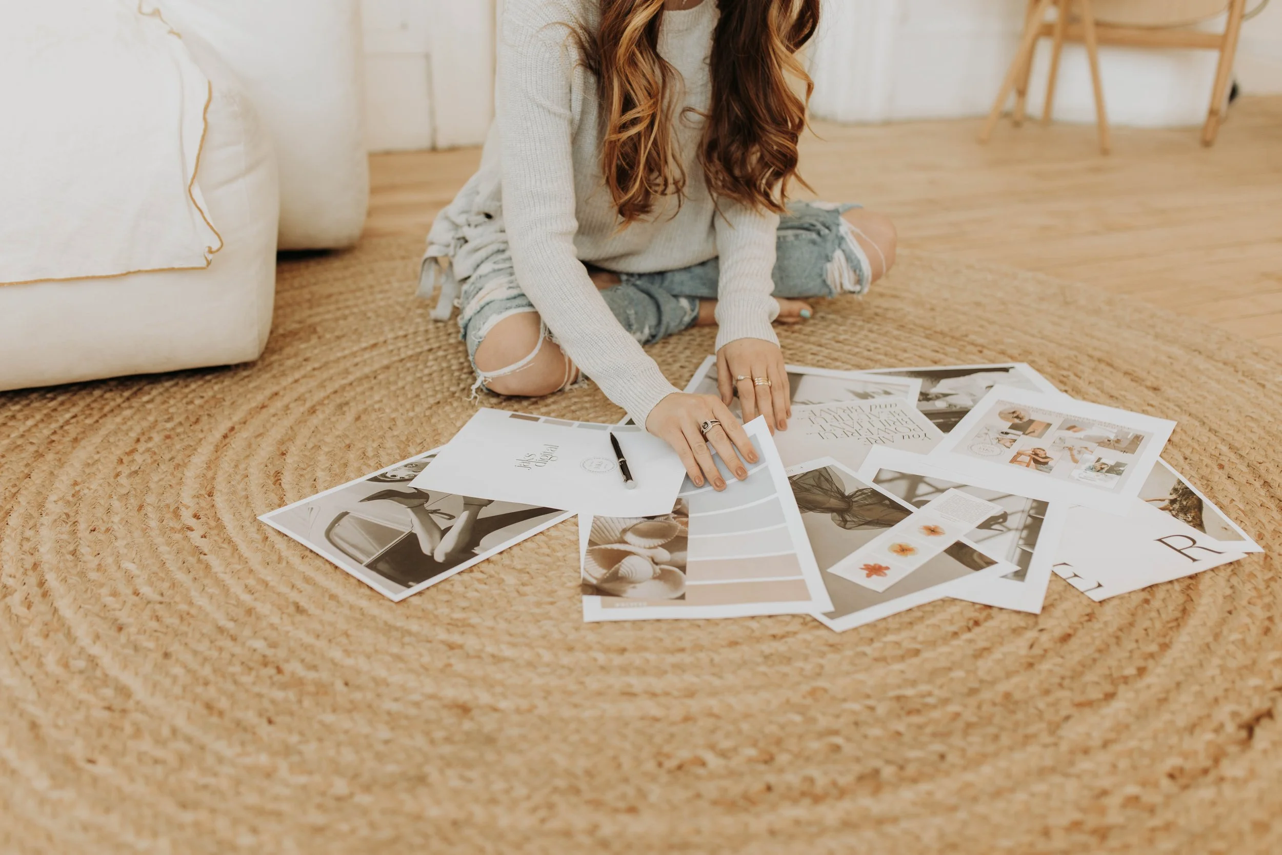 A woman is sitting cross-legged on a woven rug, sorting through printed photos and magazine pages spread out on the floor.