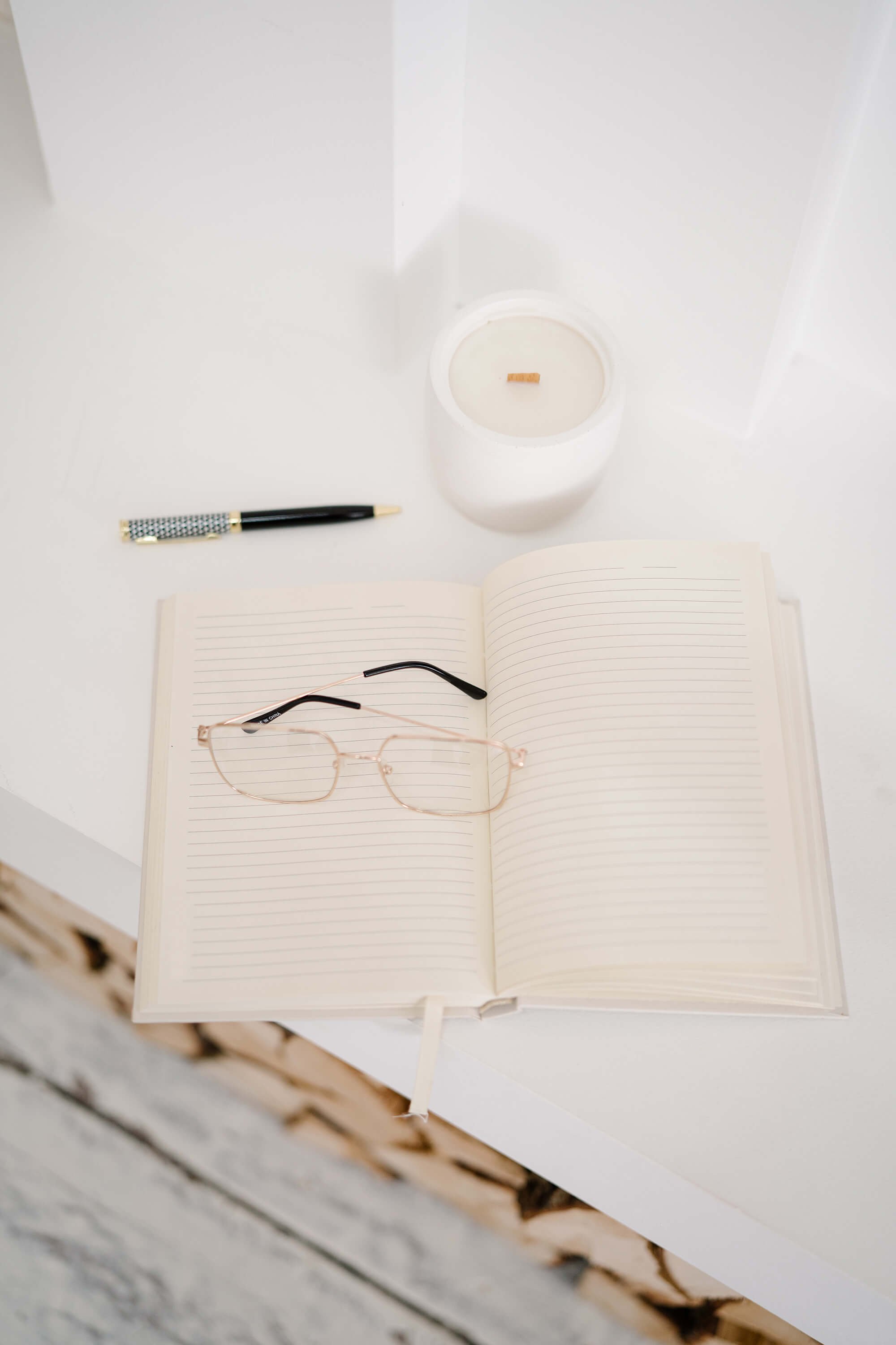 A white desk with a notebook, a pen, a pair of glasses, a lit candle, and a small box on it.