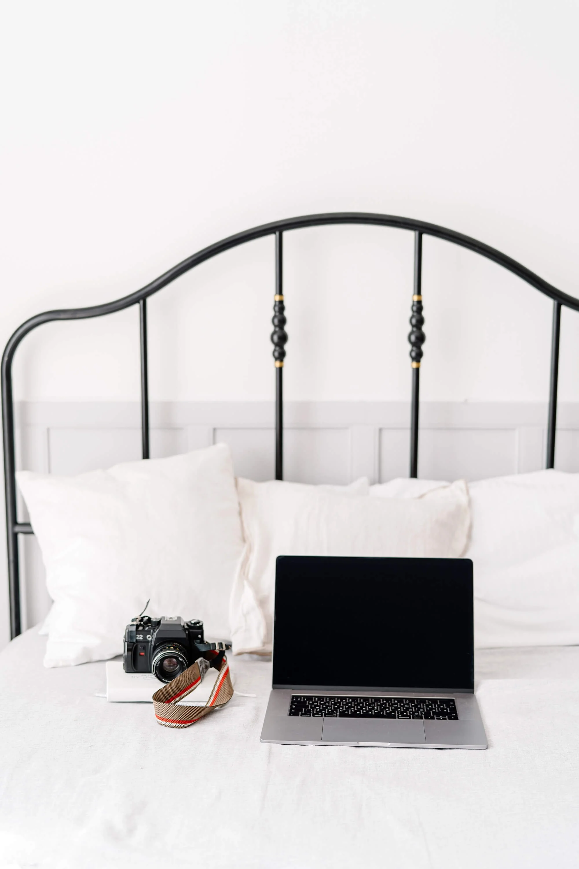 A bed with white pillows, an open laptop, a camera, and a camera strap on a white bedspread.