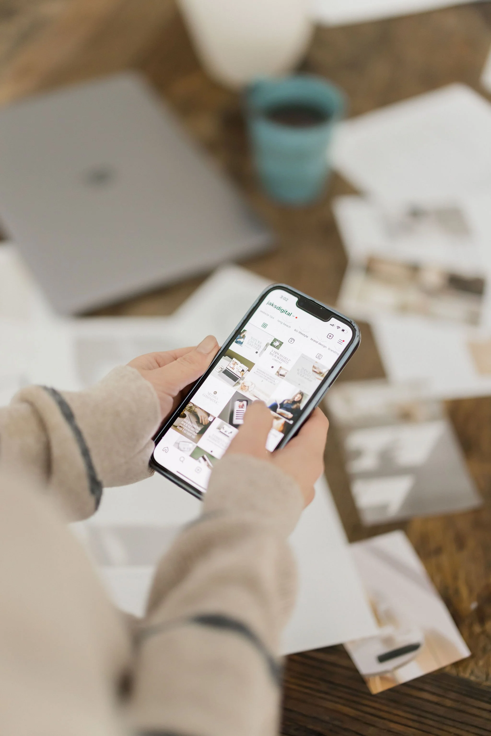 Person holding a smartphone with a social media app open, on a table with a laptop, a cup of coffee, and papers or magazines.