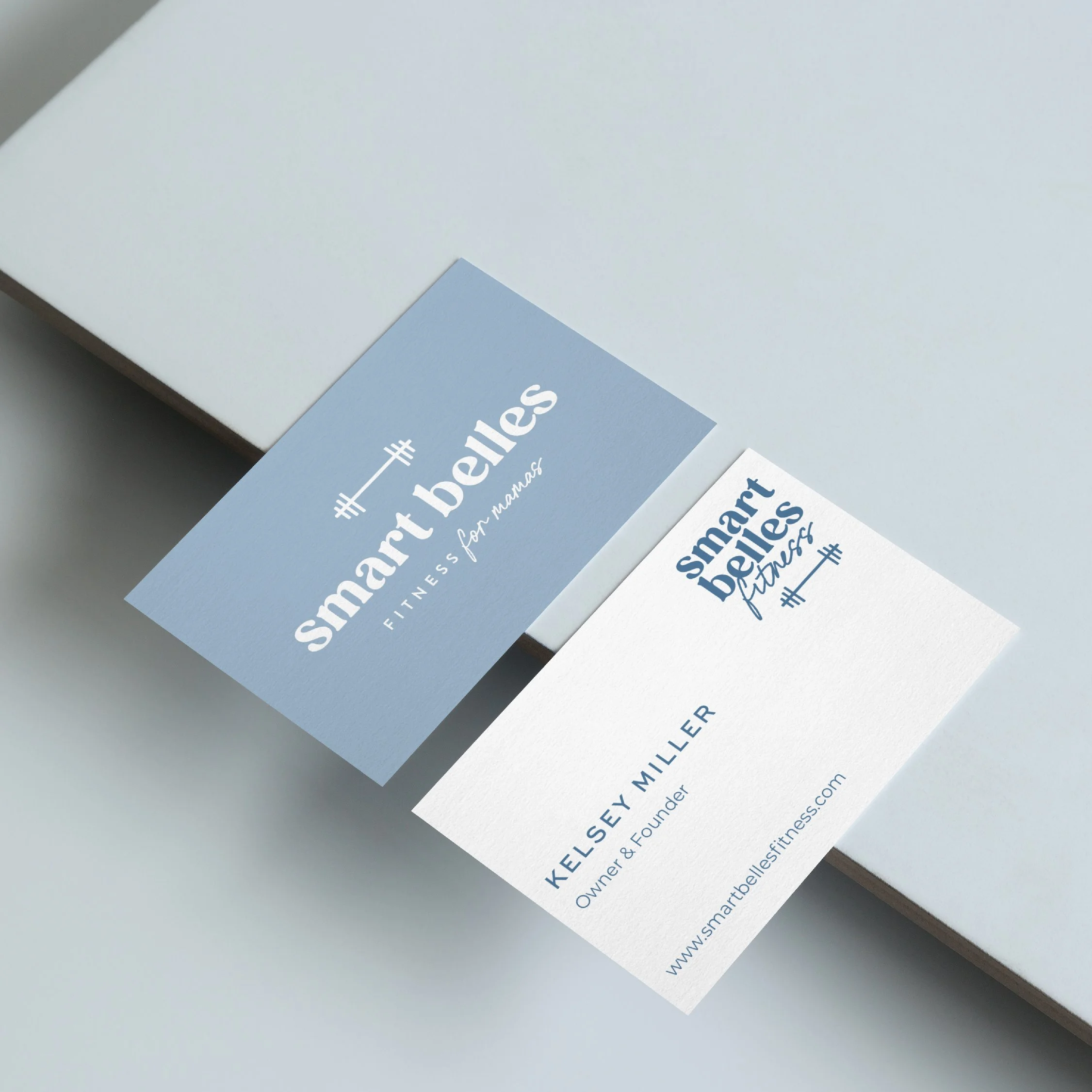 Close-up of two business cards for smart belles fitness, one on a white surface and the other partially on a gray surface. The cards display the logo, business name, tagline, owner's name, title, and website.