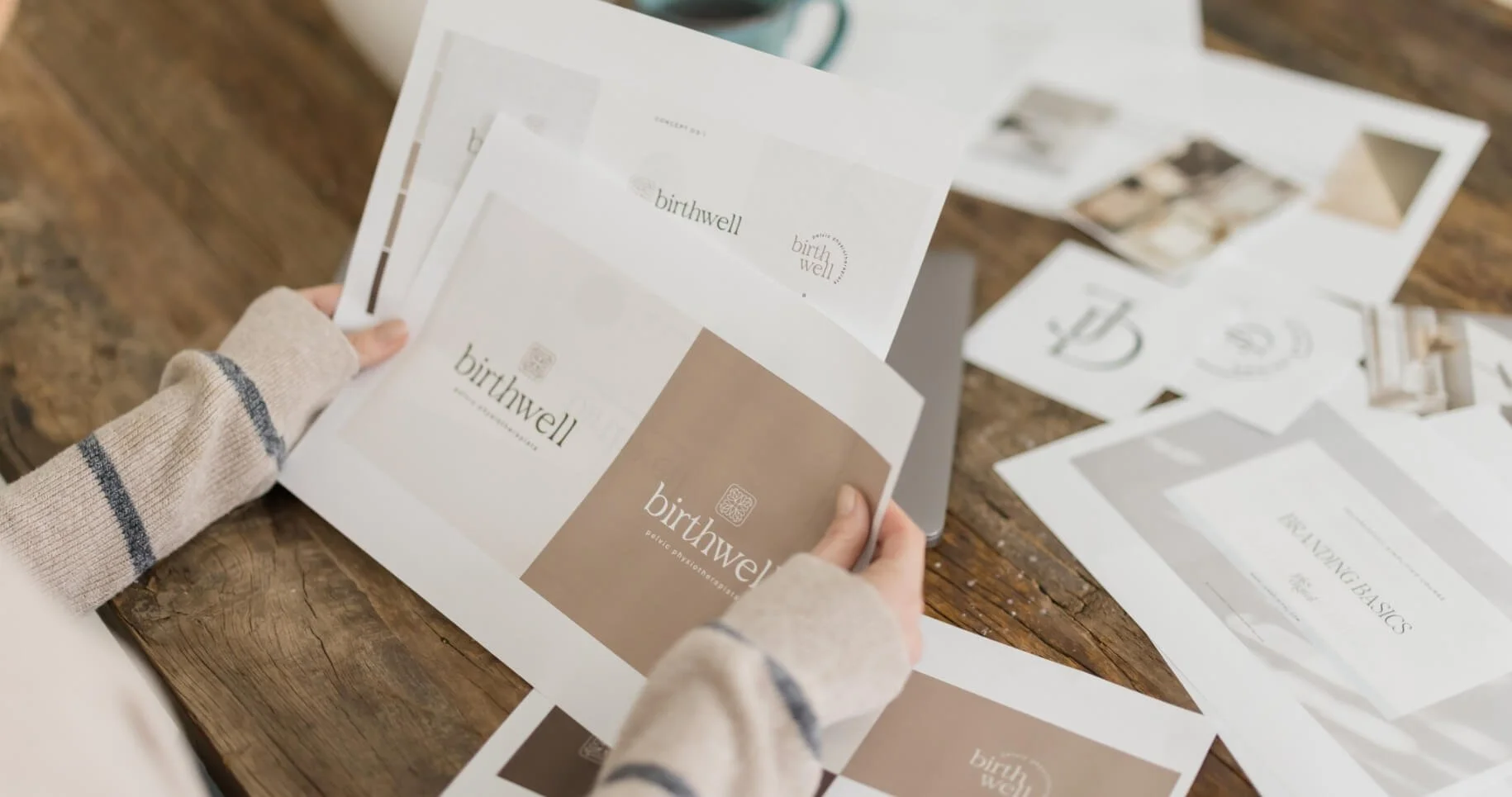 Person holding printed branding and packaging designs for a brand called 'birthwell' with various sheets of paper on a wooden table.