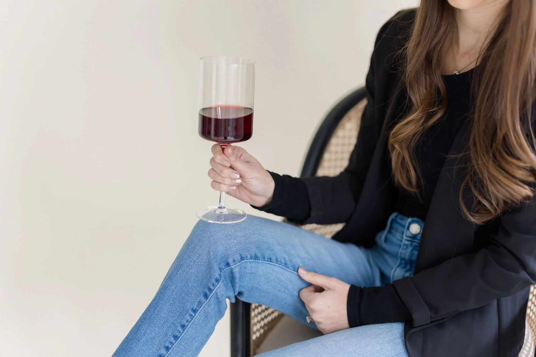 Woman sitting on a chair with one leg crossed, holding a glass of red wine in her right hand, wearing a black jacket and blue jeans.